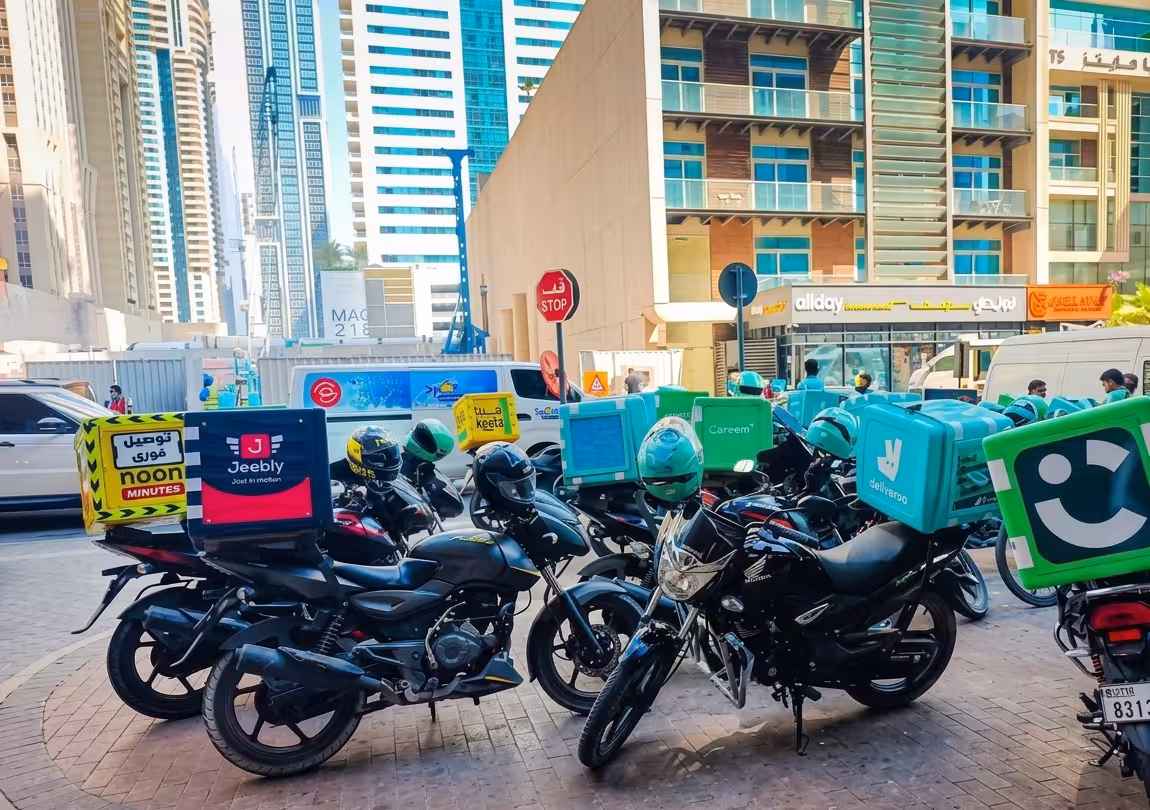 Multiple delivery company motorcycles parked on a Dubai street including Noon, Jeebly, Keeta, Deliveroo and Careem branded bikes with the city skyline in the background