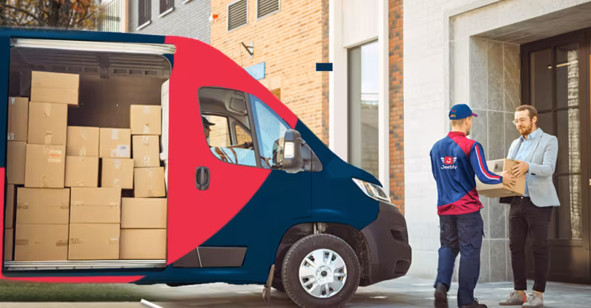 A Jeebly courier service delivery van filled with cardboard boxes parked outside a building in the UAE, while a courier uniform-clad driver hands over a parcel to a customer.