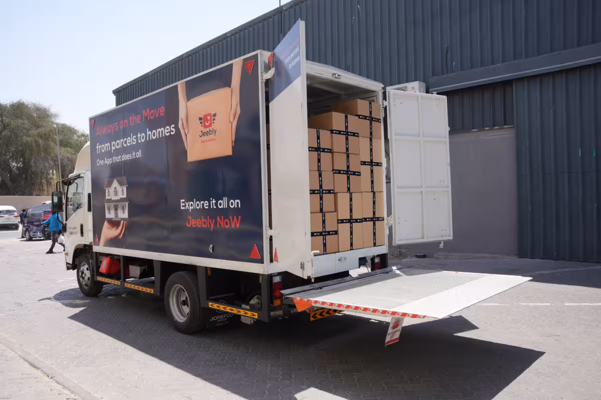 A medium-sized Jeebly delivery and moving truck parked outside a warehouse with its back doors wide open and a hydraulic lift gate lowered to the ground. Inside the cargo area, brown cardboard boxes are stacked neatly. The side of the truck features a dark blue advertisement wrap with the Jeebly logo, displaying a hand holding a house model under the text "Always on the Move from parcels to homes" and a call to action to "Explore it all on Jeebly NoW".